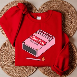 May include: Red sweatshirt with a graphic of a pink matchbox with the text "Slow Burn Book Club".