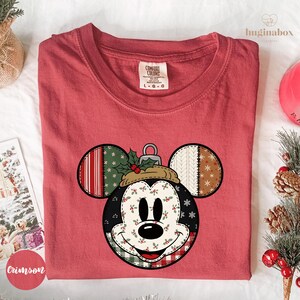May include: A red Comfort Colors t-shirt featuring a Mickey Mouse design with a Christmas ornament theme. The design includes a patchwork of patterns and colors, including red, green, white, and brown, with holly and snowflakes.