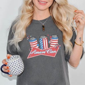 May include: Dark gray t-shirt featuring a graphic of three beverage cans with red, white, and blue designs, and the text "Ameri Can" on a red banner. The shirt is a casual style.