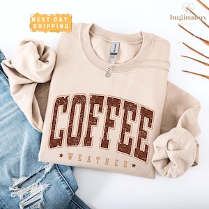 May include: A beige crewneck sweatshirt featuring the word COFFEE in large, distressed brown block letters, with the word WEATHER and two small stars printed below. The garment includes a Gildan brand tag at the inner neckline.
