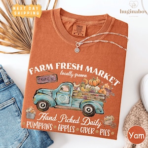 May include: An orange Comfort Colors t-shirt featuring a blue vintage truck graphic laden with pumpkins, apples, and flowers. The text reads "Farm Fresh Market" and "Hand Picked Daily". A silver necklace with a pendant is draped on the shirt.