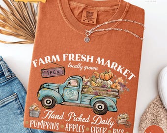 May include: An orange Comfort Colors t-shirt with a graphic of a blue vintage truck carrying pumpkins, apples, and flowers. The text reads "Farm Fresh Market" and "Hand Picked Daily". A silver necklace with a pendant is draped on the shirt.