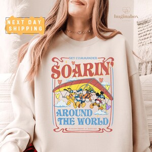 May include: Cream-colored sweatshirt featuring a graphic of Mickey Mouse, Donald Duck, Goofy, and Pluto hang gliding. The design includes the words "SOARIN'" and "AROUND THE WORLD" in a vintage style. The sweatshirt has a crew neck.