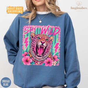 May include: An indigo blue crewneck sweatshirt featuring a graphic print of a leopard with its mouth open, wearing a silver chain necklace. The design includes the text STAY WILD in pink, surrounded by pink hibiscus flowers and neon lightning bolts.