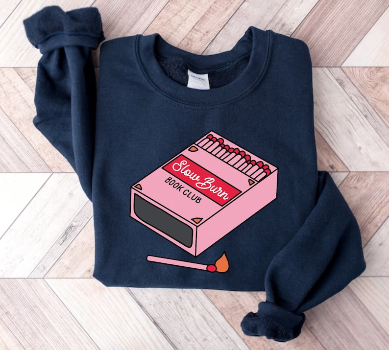 May include: A navy blue sweatshirt with a pink and red graphic of a matchbox with the text "Slow Burn Book Club".