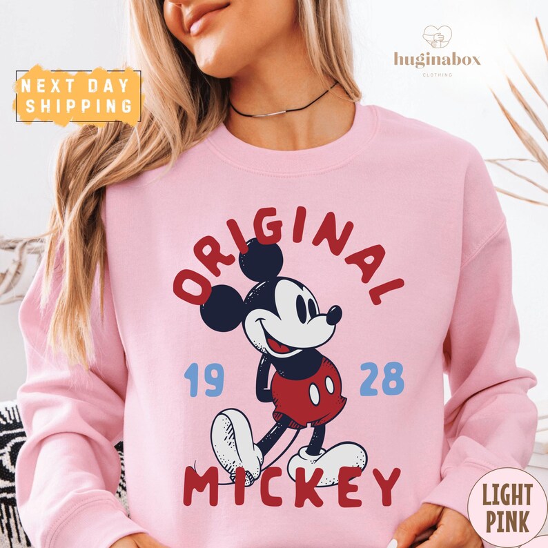 A woman wearing a pink sweatshirt with a Mickey Mouse graphic and text.