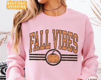 May include: A light pink crewneck sweatshirt featuring the text FALL VIBES in a distressed brown font. Below the text, a circular graphic contains a brown pumpkin illustration centred between two horizontal lines with a distressed texture.