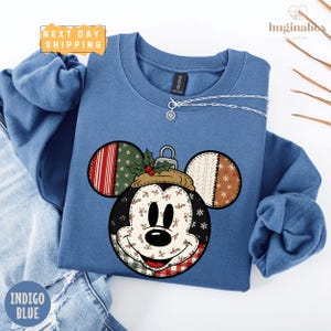 May include: Indigo blue sweatshirt featuring a Mickey Mouse ornament design. The ornament has a patchwork design with red, green, and white patterns. The sweatshirt has a crew neck and long sleeves. A silver necklace is draped across the sweatshirt.