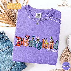 a purple t-shirt with Disney characters printed on it, along with a pair of jeans and a beige sweater.