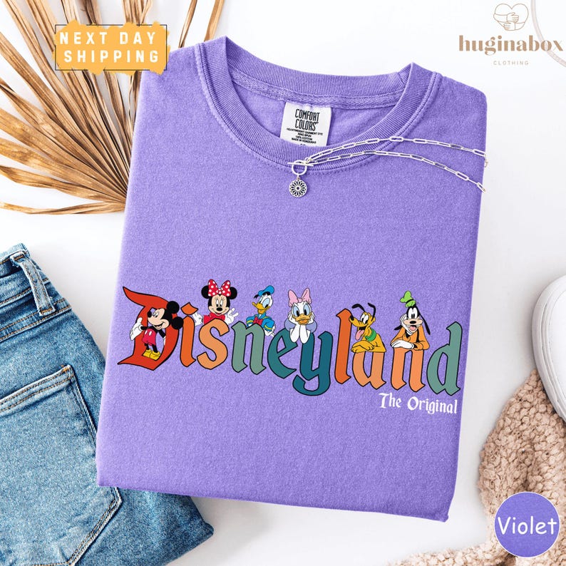a purple t-shirt with Disney characters printed on it, along with a pair of jeans and a beige sweater.