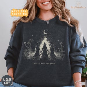 May include: A dark grey crewneck sweatshirt featuring a graphic print of three figures in pointed hats standing in a field under a crescent moon. The design includes the text "girls will be girls" in a small, simple font centered below the figures.