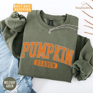 May include: An olive green sweatshirt with the words "PUMPKIN SEASON" in orange block letters. The sweatshirt has long sleeves and a crew neck. The words "NEXT DAY SHIPPING" are in a yellow box in the upper left corner. The words "MILITARY GREEN" are in a green circle in the lower left corner.