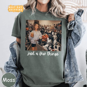 May include: A moss green t-shirt featuring a graphic of a doll holding a coffee cup in a store filled with Halloween decorations, including pumpkins and skeletons. The text "just a few things" is printed in white script below the graphic.