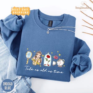 May include: Indigo blue sweatshirt featuring a graphic from Beauty and the Beast. The design includes Belle, a mirror, a rose in a glass case, and the text "Tale as old as time." A silver necklace is draped across the top.