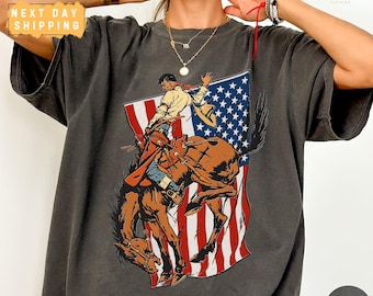 Puede incluir: Camiseta gris oscuro con un gráfico de un vaquero montando un caballo salvaje frente a una bandera estadounidense. La camiseta tiene el texto "NEXT DAY SHIPPING" en la esquina superior izquierda.