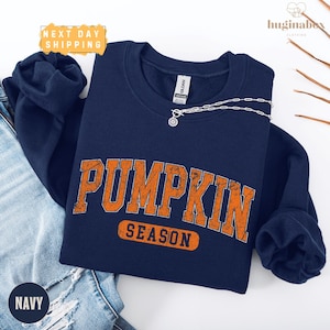 A navy blue sweatshirt with the word "PUMPKIN" printed in orange letters, along with the word "SEASON" below it, is displayed on a white background.
