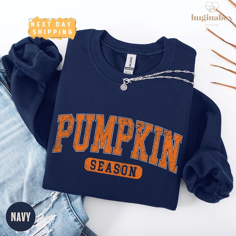A navy blue sweatshirt with the word "PUMPKIN" printed in orange letters, along with the word "SEASON" below it, is displayed on a white background.