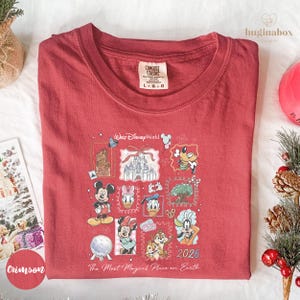 A red t-shirt with a Disney-themed graphic design, featuring various Disney characters and landmarks, such as Mickey Mouse, Minnie Mouse, Donald Duck, and the Eiffel Tower. The t-shirt is laid out on a white surface, surrounded by holiday decorations like pine cones and ornaments.