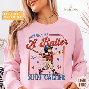 May include: Pale pink sweatshirt with a baseball graphic. The design shows a vintage illustration of a child swinging a bat, with the text "Wanna Be A Baller Shot Caller". The sweatshirt has a round neck.
