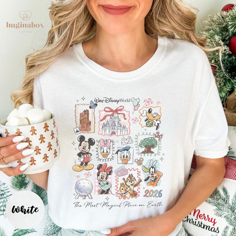 A woman wearing a white t-shirt with Disney characters and a mug of marshmallows, smiling and holding a Christmas tree.