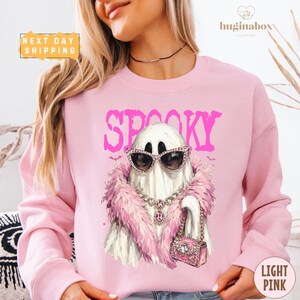 May include: A light pink crewneck sweatshirt featuring a graphic of a white ghost wearing pink-rimmed sunglasses, a pink feather boa, a jeweled necklace, and carrying a pink handbag. The word SPOOKY is printed in large pink letters above the ghost.
