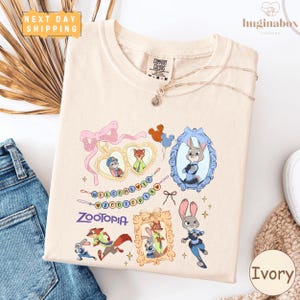 a white t-shirt with a colorful graphic design of cartoon animals, including a rabbit, fox, and other animals, along with a welcome message and the word "Zooper". The t-shirt is displayed on a white background, and there are some jeans and a white object visible in the background.
