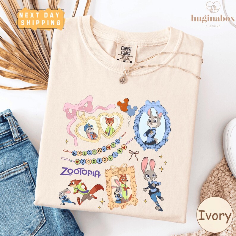 a white t-shirt with a colorful graphic design of cartoon animals, including a rabbit, fox, and other animals, along with a welcome message and the word "Zooper". The t-shirt is displayed on a white background, and there are some jeans and a white object visible in the background.
