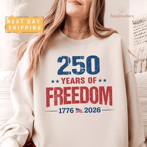 May include: Cream-colored sweatshirt with the text "250 YEARS OF FREEDOM" in blue and red. The design includes stars, the years 1776 and 2026, and a small American flag graphic. The sweatshirt has a crew neck.