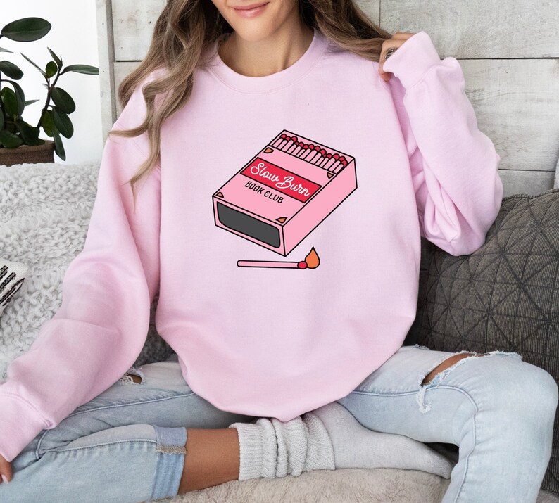 May include: A pink sweatshirt with a graphic of a matchbox that says "Slow Burn Book Club" and a lit match below it.