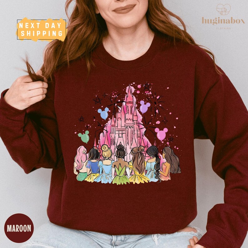 A woman wearing a burgundy sweatshirt with a colorful illustration of a group of women in various outfits standing in front of a castle.