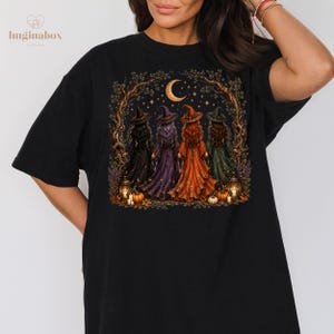 A woman wearing a black t-shirt with a graphic design depicting four witch figures in a nighttime scene with a crescent moon and stars.