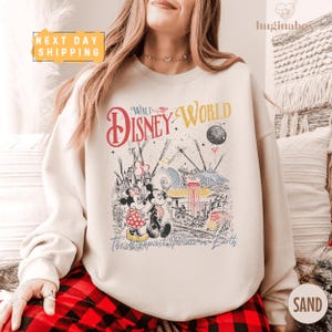 A woman wearing a white sweatshirt with a Disney World-themed graphic printed on it, sitting on a couch.