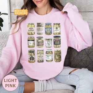 a woman sitting on a couch wearing a pink sweatshirt with a jar of pickles