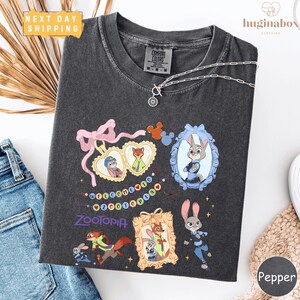a black t-shirt with a colorful graphic design of cartoon characters, including a rabbit, foxes, and other animals, along with a heart-shaped ribbon and a "Welcome to Zootopia" banner. The t-shirt is displayed on a white background, and there are some jeans and a necklace visible in the background.