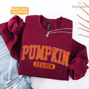 a maroon sweatshirt with the word "PUMPKIN" printed on it, along with the word "SEASON" below it. The sweatshirt is placed on a white background, and there is a pair of jeans visible in the background.
