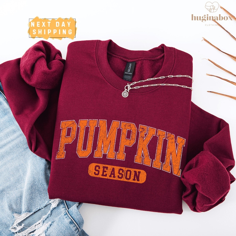 a maroon sweatshirt with the word "PUMPKIN" printed on it, along with the word "SEASON" below it. The sweatshirt is placed on a white background, and there is a pair of jeans visible in the background.