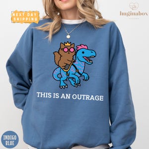 May include: Indigo blue sweatshirt featuring a cartoon cat wearing sunglasses and a crown riding a blue dinosaur. The text "THIS IS AN OUTRAGE" is printed below the graphic. The sweatshirt has a crew neck.