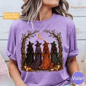 A purple t-shirt with a graphic design depicting four women dressed as witches, surrounded by autumn foliage and pumpkins, with a crescent moon and stars in the night sky.
