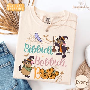 May include: Ivory-colored t-shirt with a whimsical Halloween design. The graphic features cartoon mice in witch hats, a high-heeled shoe, bats, spiderwebs, and the words "Bibbidi Bobbidi Boo" in various colors. The shirt has a Comfort Colors tag.