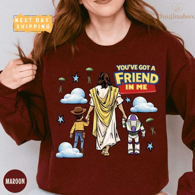 A woman wearing a maroon sweater with a graphic design depicting a religious figure holding a child and a toy robot, along with the text "You've got a Friend in Me".