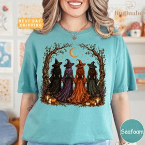 A woman wearing a teal t-shirt with a graphic design depicting four women dressed in witch costumes standing in a circle under a crescent moon.