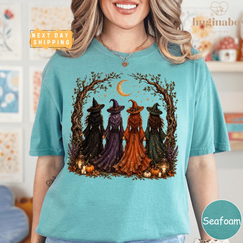 A woman wearing a teal t-shirt with a graphic design depicting four women dressed in witch costumes standing in a circle under a crescent moon.