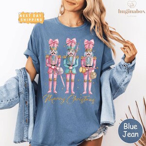 Puede incluir: Camiseta azul vaquero con una ilustración en acuarela de tres soldados cascanueces. Los soldados visten uniformes rosas y verde azulado con lazos rosas. El texto "Merry Christmas" está en escritura dorada. La camiseta tiene el texto "Blue Jean" en la parte inferior derecha.