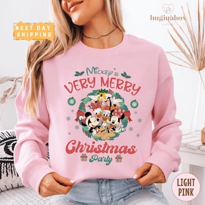 May include: Light pink sweatshirt featuring Mickey Mouse and friends with the text "Mickey's Very Merry Christmas Party". The design includes a wreath, snowflakes, and Christmas gifts. The sweatshirt has long sleeves.