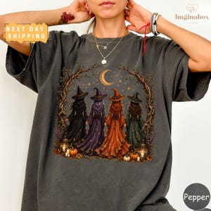 A person wearing a gray t-shirt with a graphic design depicting four women dressed in witch costumes, surrounded by autumn elements such as pumpkins and a crescent moon.
