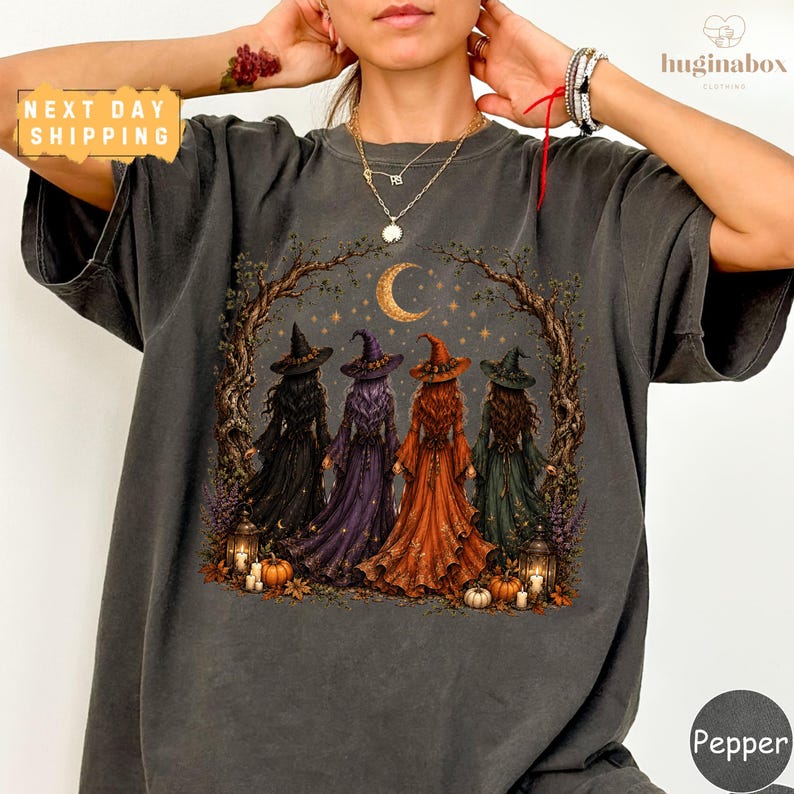 A person wearing a gray t-shirt with a graphic design depicting four women dressed in witch costumes, surrounded by autumn elements such as pumpkins and a crescent moon.