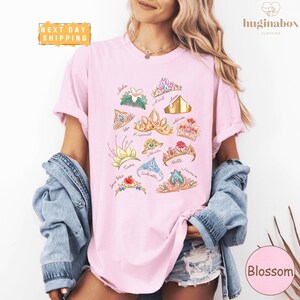 A woman wearing a pink t-shirt with a graphic design of various crowns and floral elements, paired with denim clothing and a denim jacket.