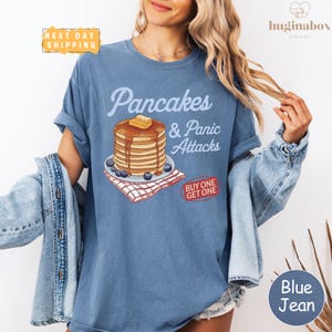May include: Blue jean t-shirt with the text "Pancakes & Panic Attacks" and a graphic of pancakes with butter and syrup. The shirt also has a "Buy One Get One" promotion.