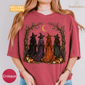 A woman wearing a pink t-shirt with a graphic design depicting four women dressed as witches, surrounded by autumnal elements such as pumpkins and a crescent moon.