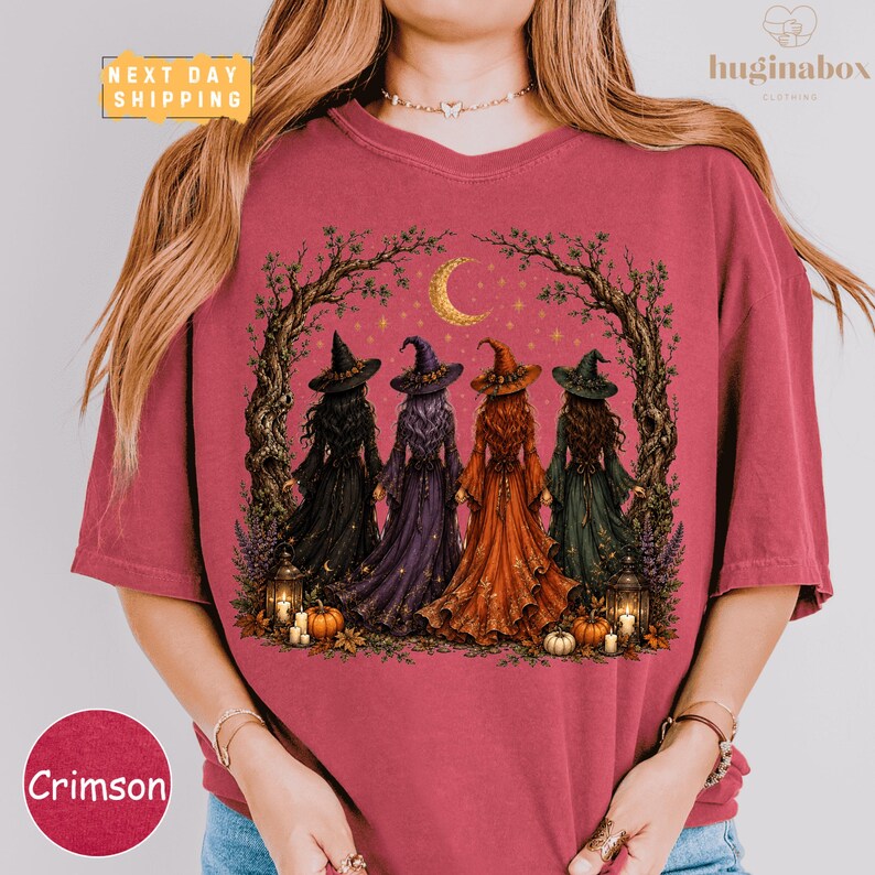 A woman wearing a pink t-shirt with a graphic design depicting four women dressed as witches, surrounded by autumnal elements such as pumpkins and a crescent moon.